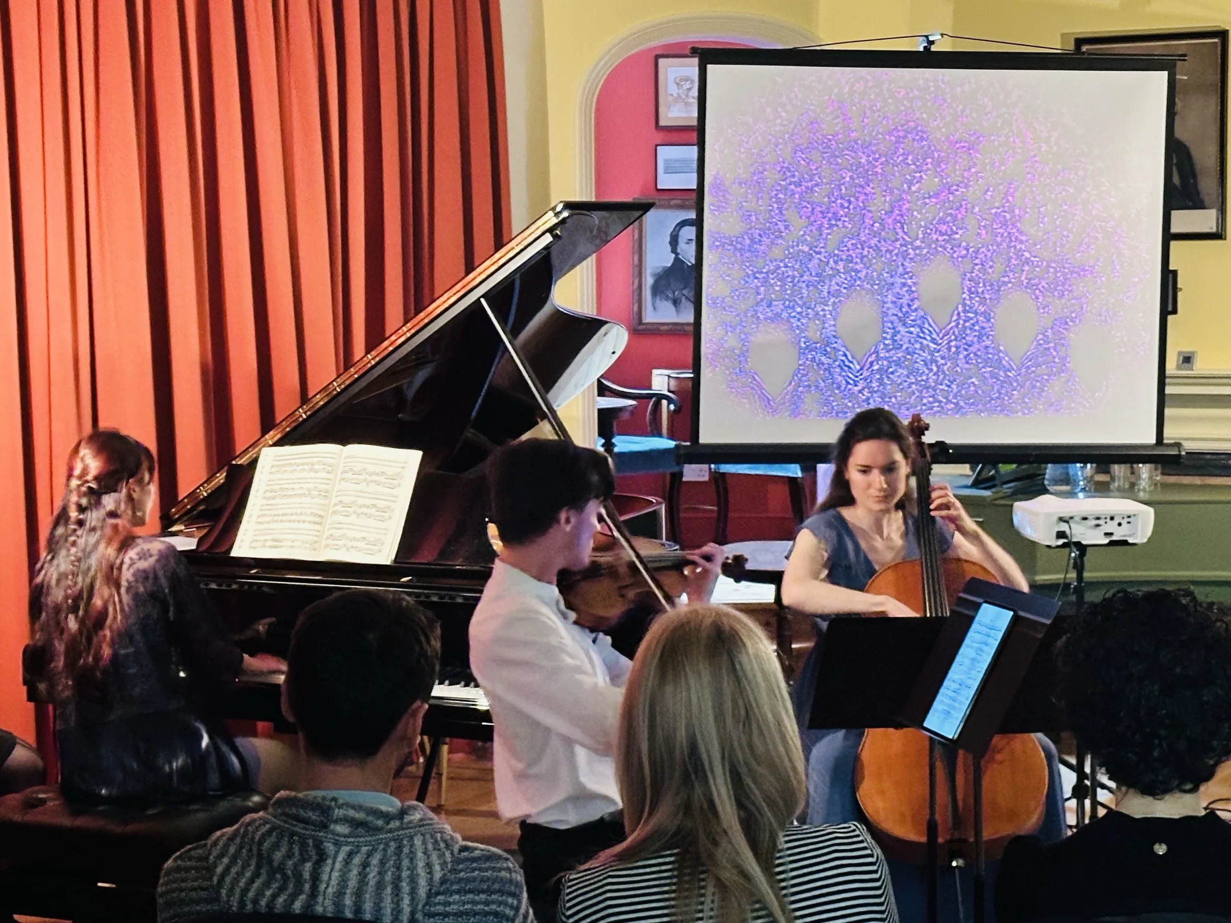A piano trio performs beside a projection screen in a red-curtained hall